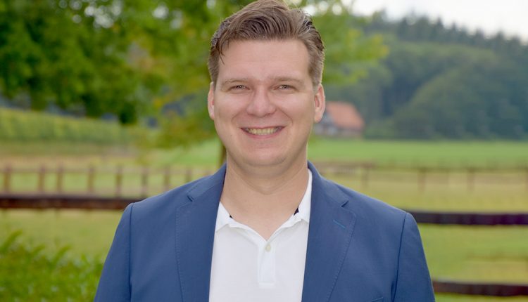 Man in a suit standing in front of a field with a fence.