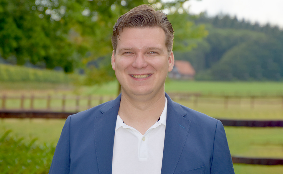 Man in a suit standing in front of a field with a fence.