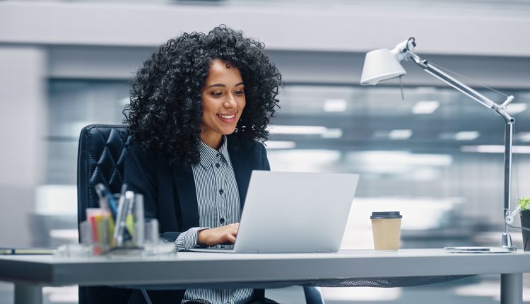 Modern Office: Black Businesswoman Sitting at Her Desk Working on a Laptop Computer. Smiling Successful African American Woman working with Big Data e-Commerce. Motion Blur Background