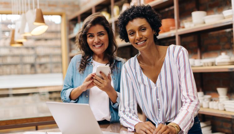 Happy ceramists smiling at the camera while working with wireless technology. Female entrepreneurs managing online orders in their store. Two businesswomen running a creative small business together.