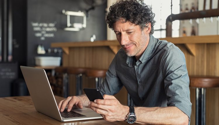 Happy mature business man sitting at cafeteria with laptop and smartphone. Businessman texting on smart phone while sitting in a pub restaurant. Portrait of senior formal man working and checking email on computer.