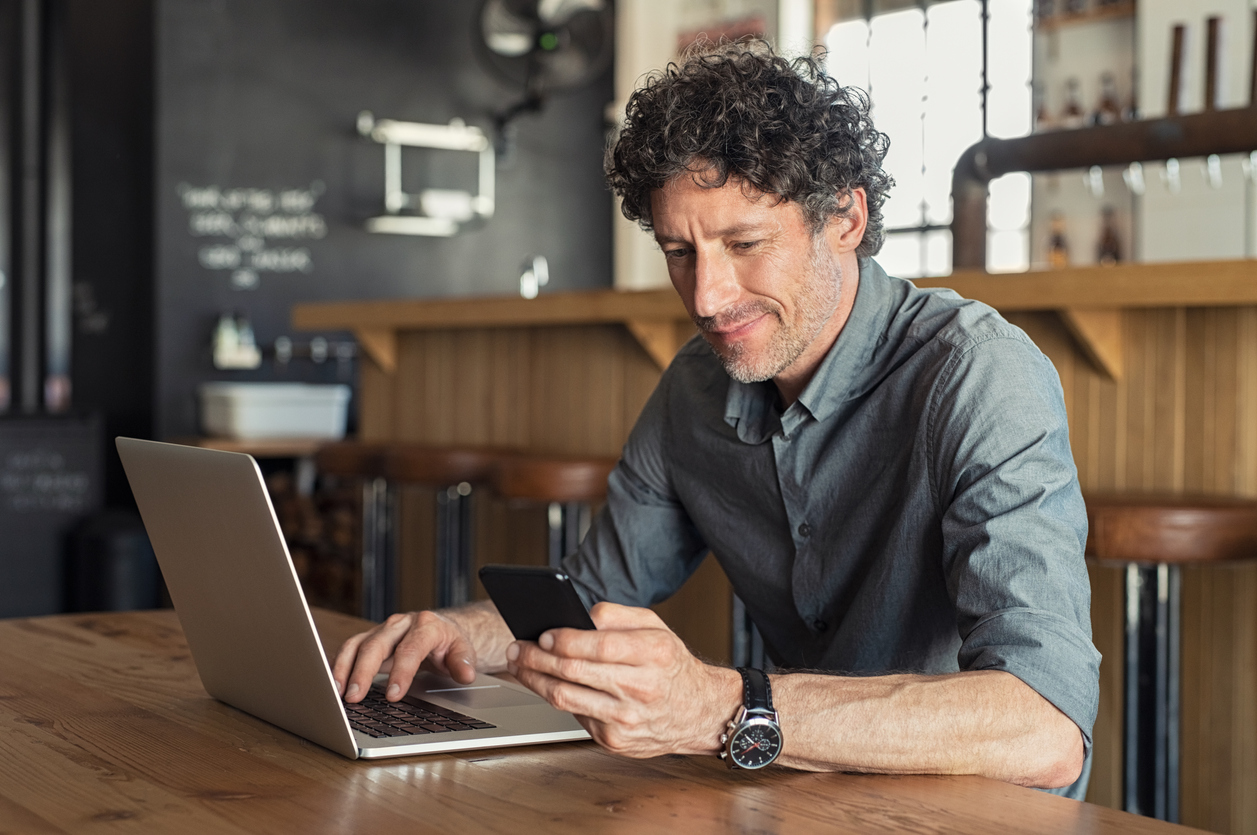 Happy mature business man sitting at cafeteria with laptop and smartphone. Businessman texting on smart phone while sitting in a pub restaurant. Portrait of senior formal man working and checking email on computer.