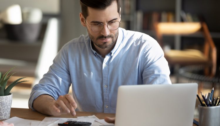 Focused young Caucasian man look at laptop screen calculate expenses expenditures pay bills taxes online. Millennial male busy managing household family budget, take care of financial paperwork.