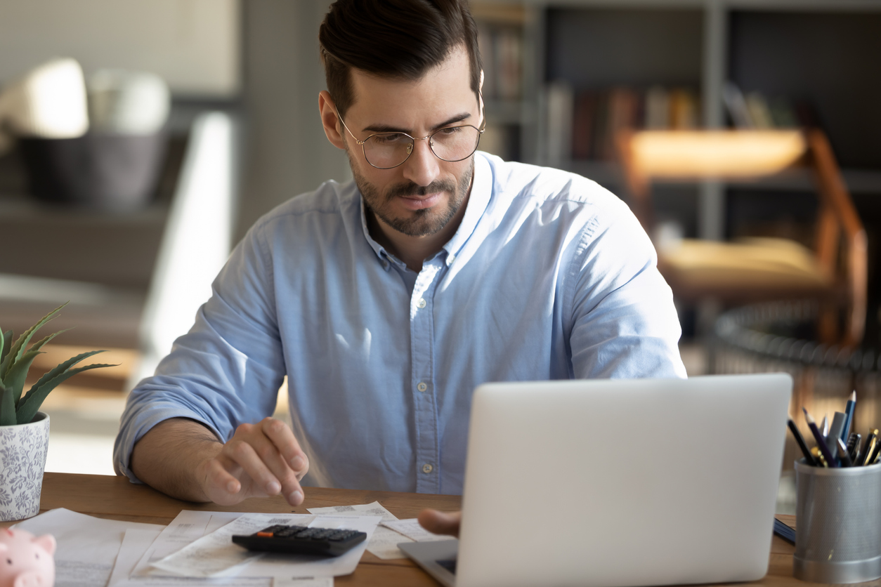 Focused young Caucasian man look at laptop screen calculate expenses expenditures pay bills taxes online. Millennial male busy managing household family budget, take care of financial paperwork.