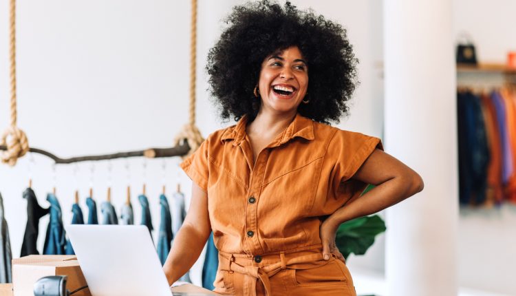 Ethnic small business owner smiling cheerfully while standing in her shop. Happy businesswoman managing her clothing orders on a laptop. Black female entrepreneur running an online clothing store.
