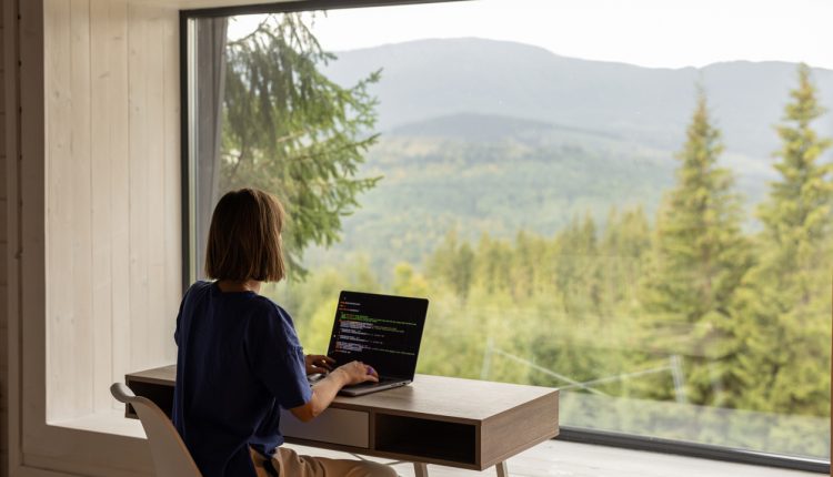 Woman works on laptop while sitting by the table in front of panoramic window with great view on mountains. Remote work and escaping to nature concept