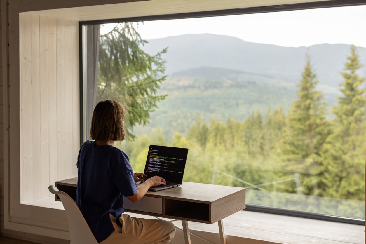 Woman works on laptop while sitting by the table in front of panoramic window with great view on mountains. Remote work and escaping to nature concept