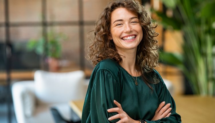Portrait of young smiling woman looking at camera with crossed arms. Happy girl standing in creative office. Successful businesswoman standing in office with copy space.