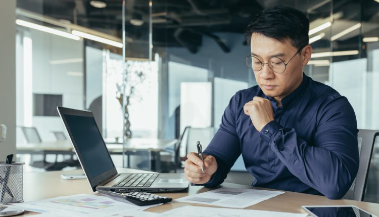 Thinking asian financier in glasses accountant working with documents and accounts, using laptop, male businessman doing paperwork in sitting at desk inside office building.