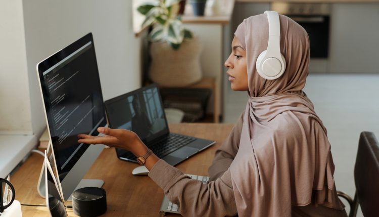 Young Muslim female programmer pointing at data on computer screen while sitting by workplace and communicating with colleague in video chat