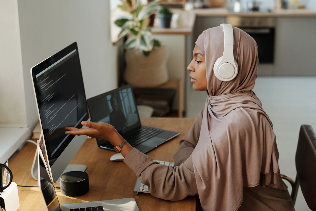 Young Muslim female programmer pointing at data on computer screen while sitting by workplace and communicating with colleague in video chat