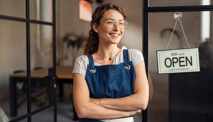 Portrait of happy waitress standing at restaurant entrance while looking outdoor. Portrait of young business woman attend new customers in her coffee shop. Smiling small business owner showing open sign in her shop while leaning against the door.