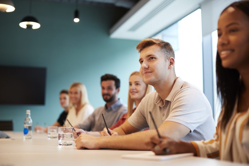 Group Of Young Candidates Sitting At Boardroom Table Listening To Presentation At Business Graduate Recruitment Assessment Day