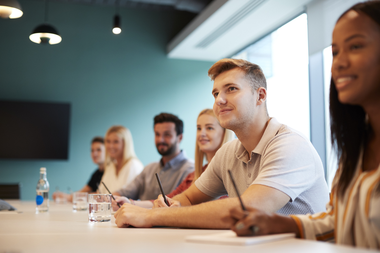 Group Of Young Candidates Sitting At Boardroom Table Listening To Presentation At Business Graduate Recruitment Assessment Day