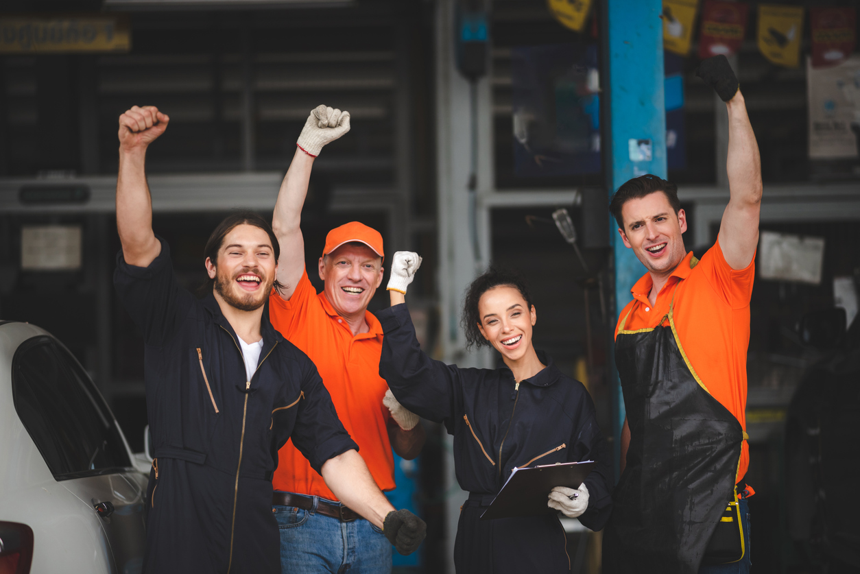 group of young and senior male and female car mechanics in garage wearing uniform enjoying and celebrating with female worker holding clipping board