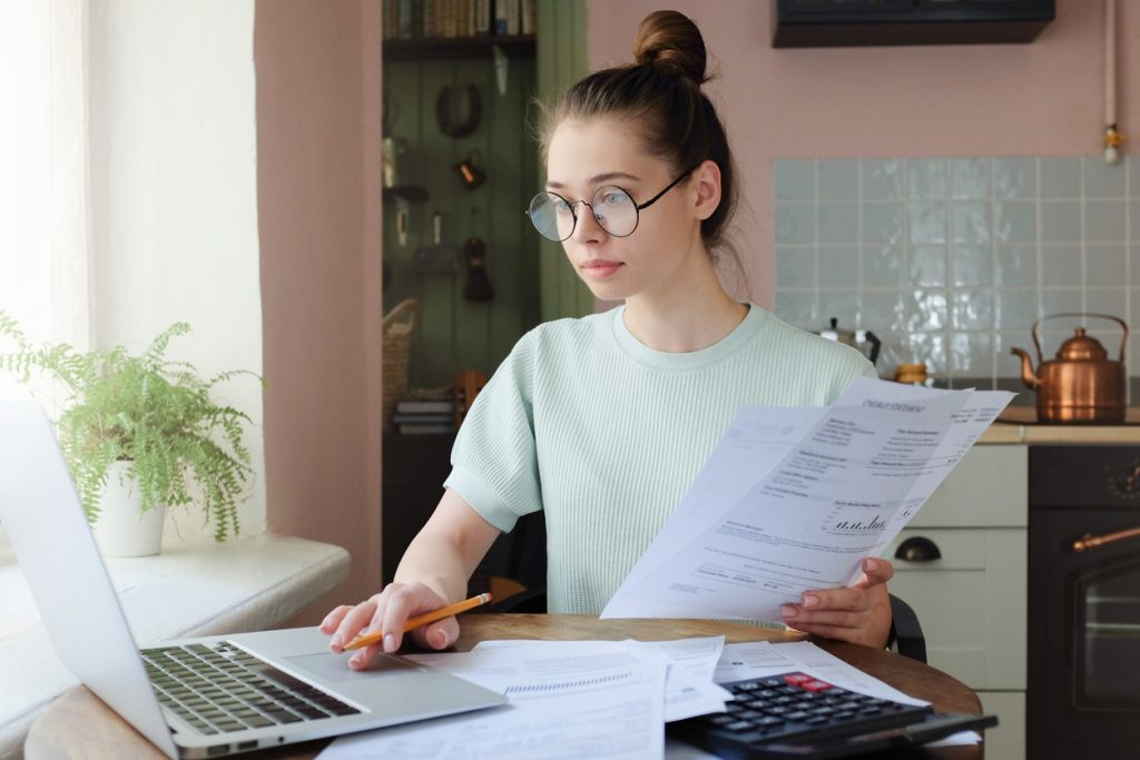 young woman managing domestic budget, sitting at kitchen table with open laptop, documents and calculator, using touchpad, making notes with pencil