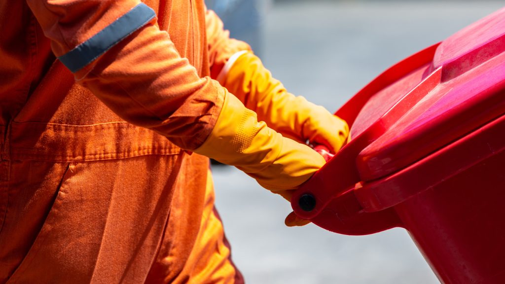 man in protective suit and disposal container for infectious waste, infectious waste must be disposed in the trash red bag, coronavirus protection equipment in medical waste bin.