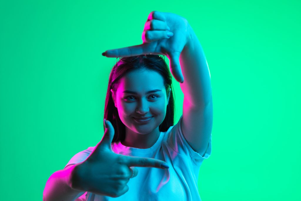 close up portrait of young pretty smiling caucasian girl showing frame gesture isolated on green background in neon light.