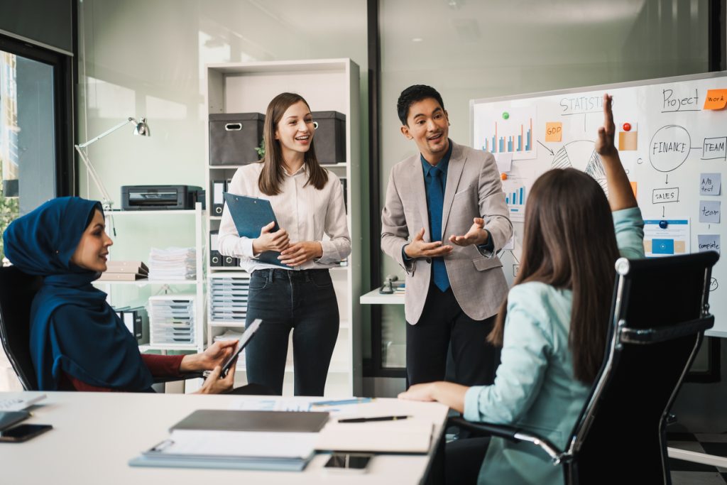 a diverse team, including a middle aged asian businessman, caucasian young businesswoman, muslim hijab wearing middle aged businesswoman, meets in a boardroom to close an annual general meeting.