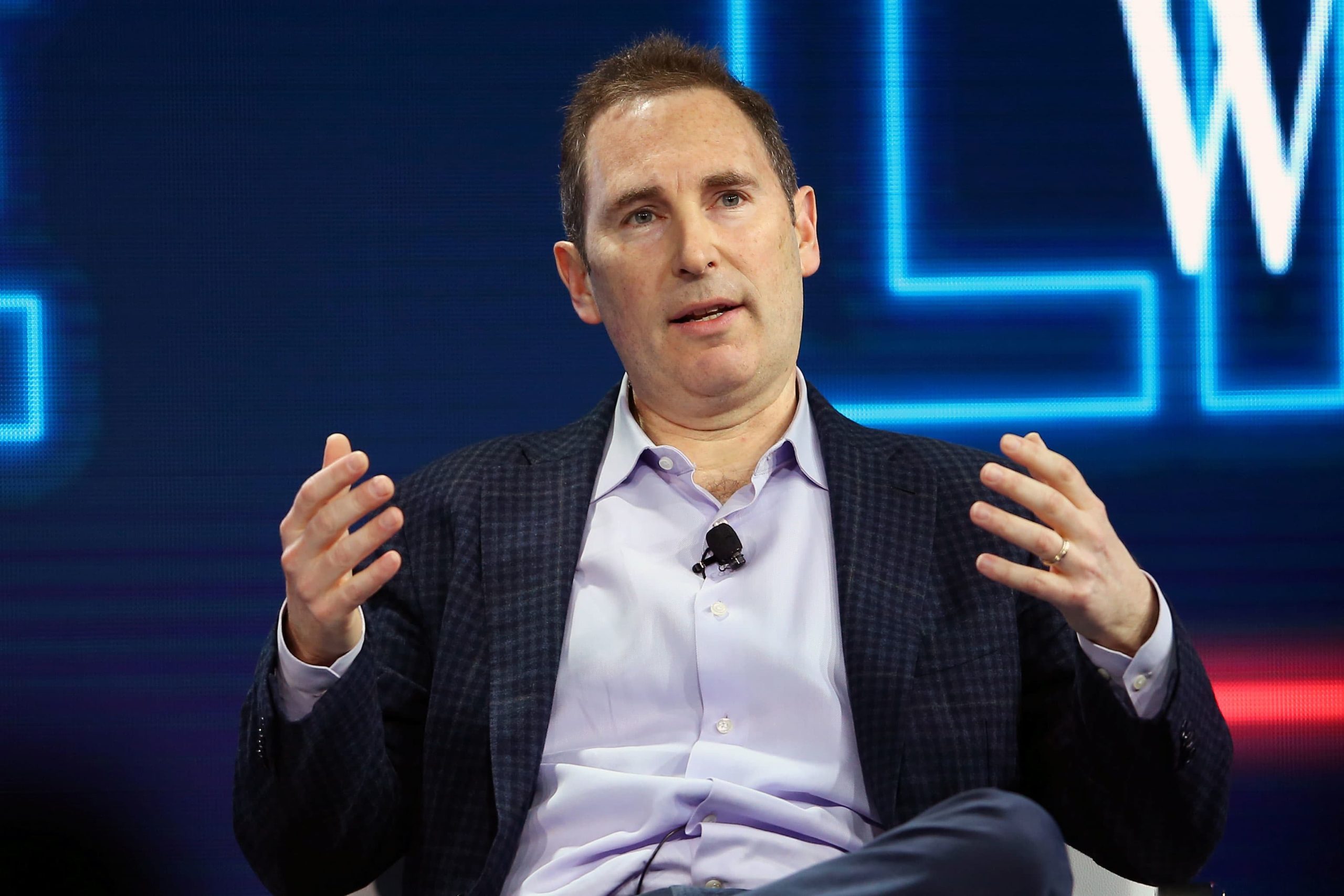 Andy Jassy speaking on stage at a tech conference, wearing a checked blazer and open-collar shirt, gesturing with both hands during a panel discussion.