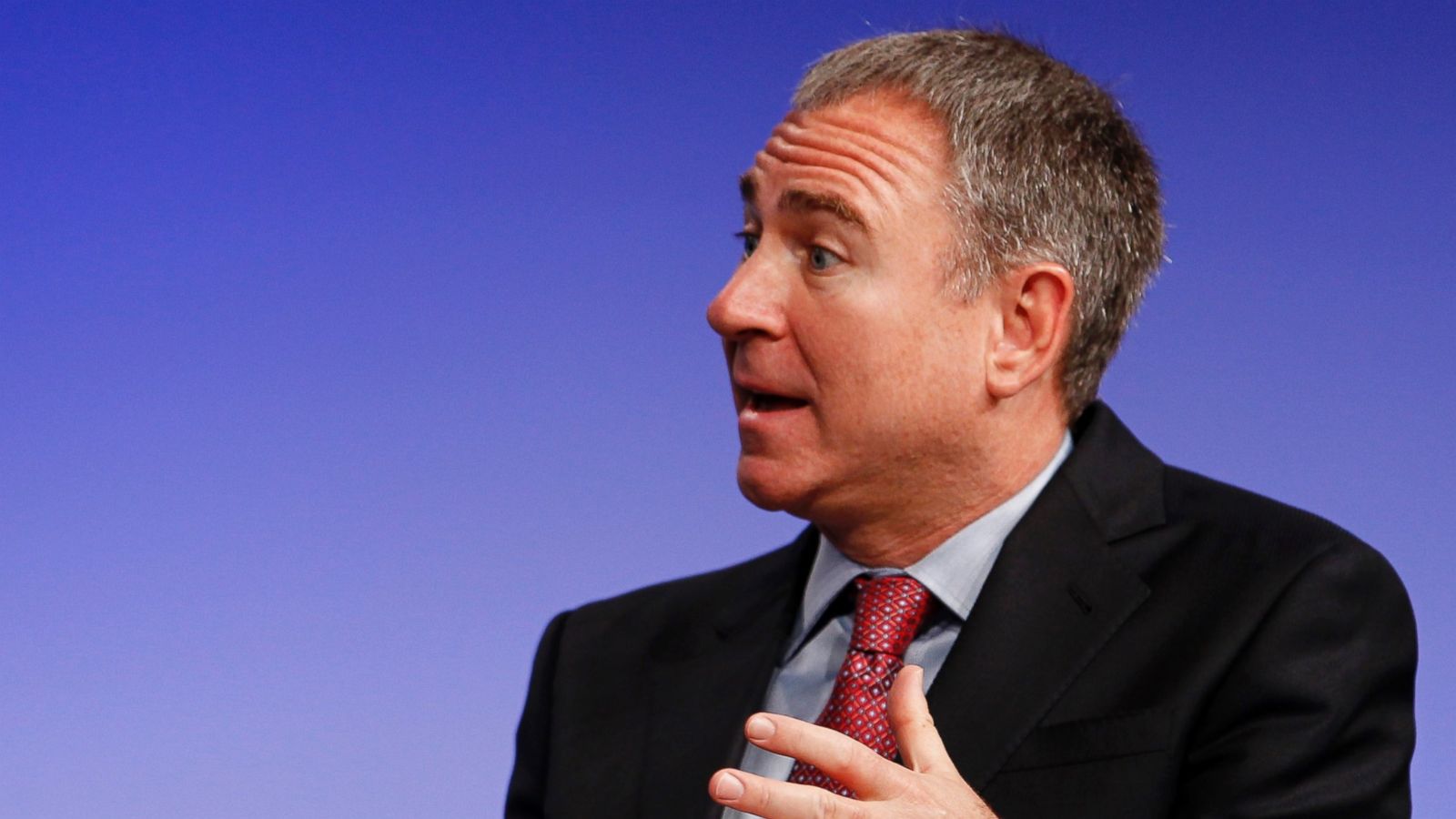 Ken Griffin speaking on stage in a dark suit and red tie, gesturing with his hand against a blue backdrop.