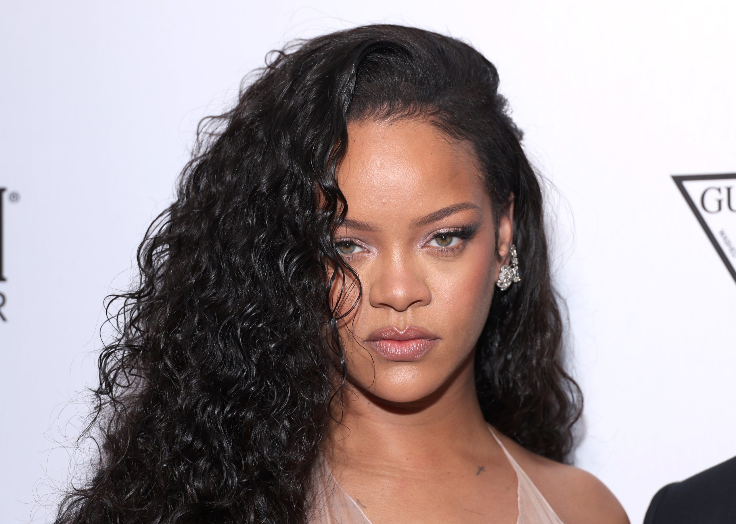 Rihanna poses for a close-up photo at a formal event, wearing a sheer nude-colored dress and diamond earrings. Her long, curly black hair is swept to one side, and she gazes directly into the camera with a confident expression.