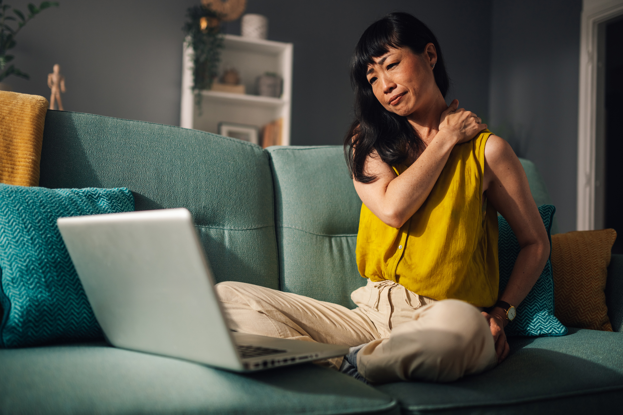 asian woman sitting on couch, holding shoulder in discomfort, laptop beside