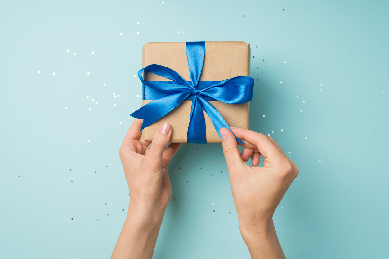 first person top view photo of hands unpacking craft paper giftbox with vivid blue satin ribbon bow over shiny sequins on isolated pastel blue background