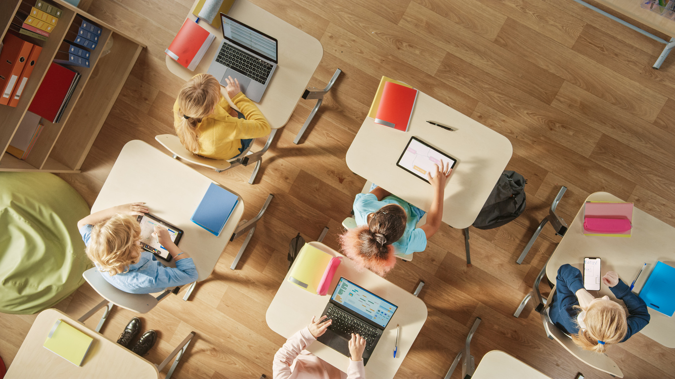 top view shot in elementary school computer science classroom: children sitting at their school desk using personal computers and digital tablets for assignments.