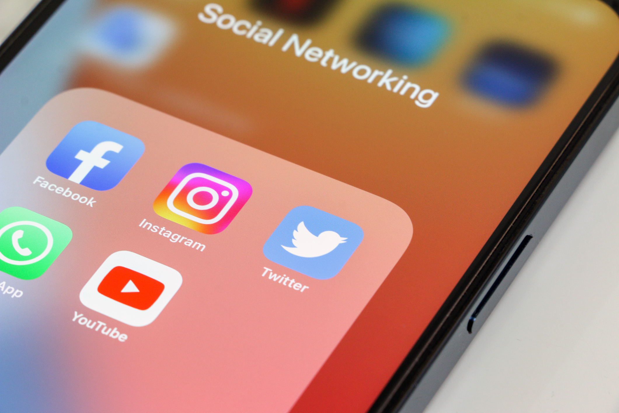 close up of popular social media app icons, facebook, instagram, twitter, whatsapp and youtube, in that order, displayed on smartphone screen with orange wallpaper. device is on white background.
