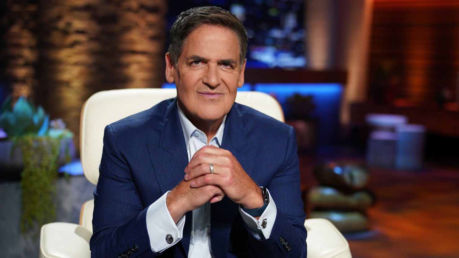 Mark Cuban sitting on the Shark Tank set, wearing a navy blue suit and clasping his hands, during a television interview segment about wealth and business.