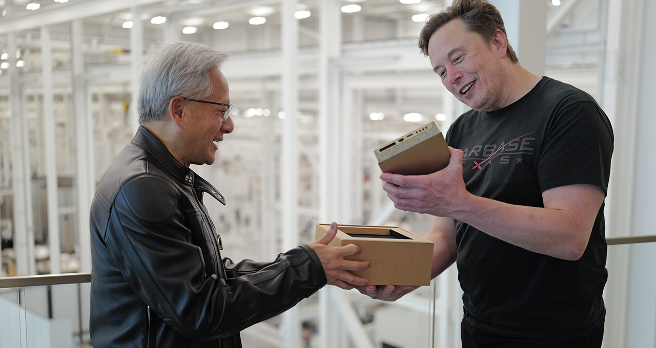 Elon Musk and NVIDIA CEO Jensen Huang smiling as they exchange a small tech device inside a modern factory setting.