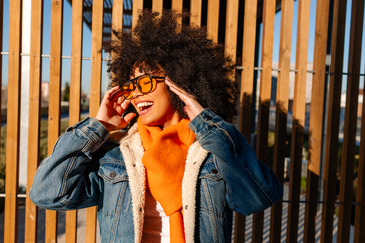 fashionable afro american woman smiling and touching sunglasses in urban setting