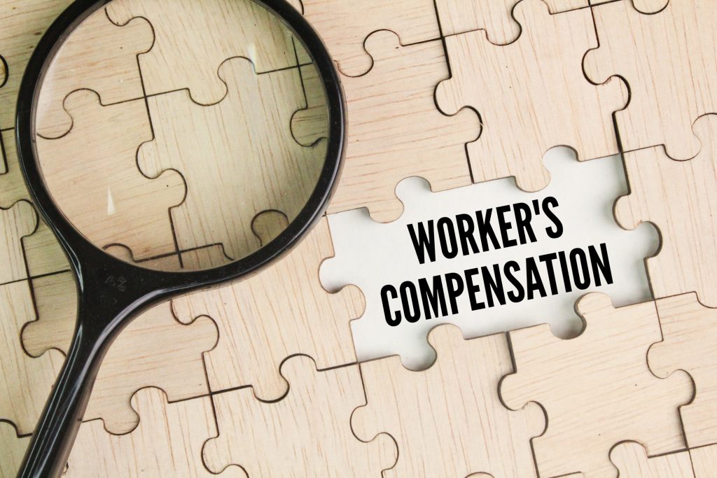 magnifying glass and wooden puzzle with the word workers' compensation. form of insurance providing wage replacement and medical benefits to injured employees