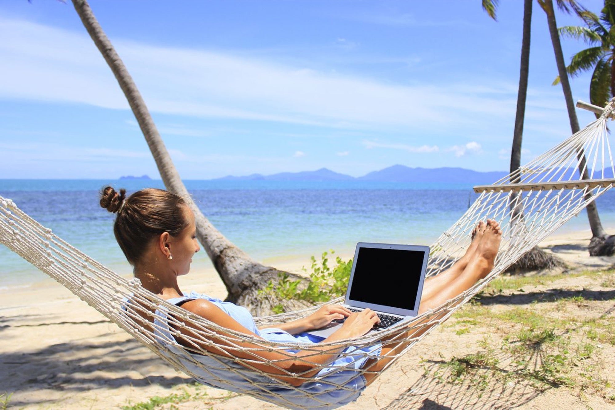 Professional working on a laptop while relaxing in a hammock on a sunny beach, representing passive income and flexible work