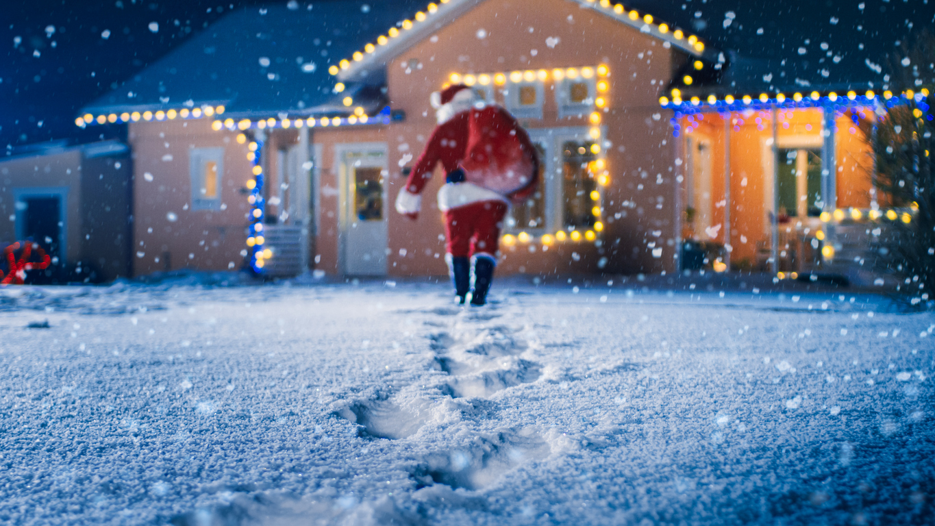 low angle shot of santa claus with red bag, walks into front yard of the idyllic house decorated with lights and garlands. santa bringing gifts and presents at night. magical new year's eve with falling snow.