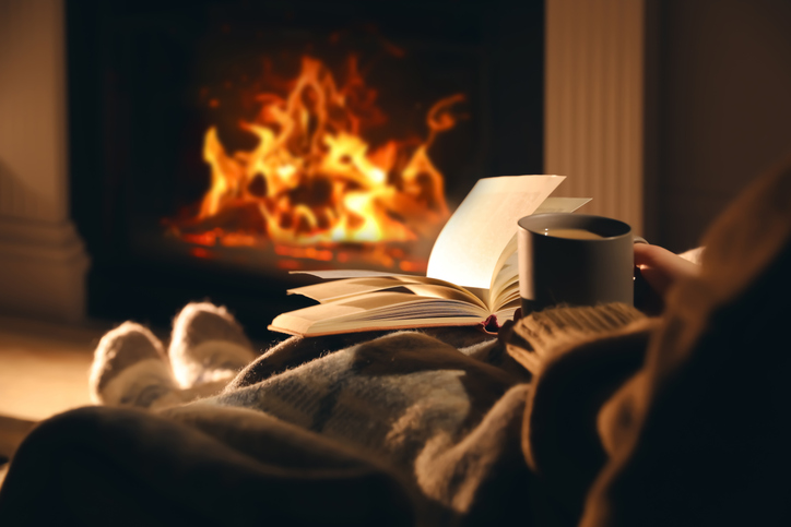 woman with cup of drink and book near fireplace at home, closeup