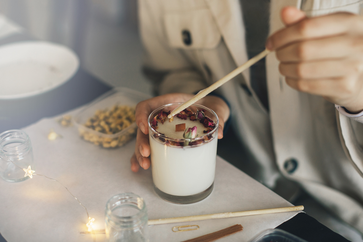 woman making candle of soy wax, decorating it with dry roses and flowers using wooden stick.