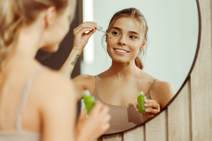young woman holding green bottle applying serum to face, looking in mirror, standing in bathroom