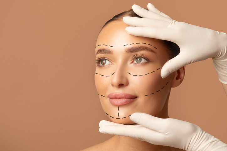 portrait of young woman and plastic surgeon making marks on female face over beige studio background, preparing for facelifting procedure