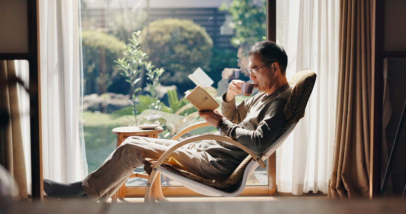 asian man, tea and relax with book on chair for reading, story or novel by window at home. japan, male person or reader with drink, beverage or chapter for fantasy, vocabulary or knowledge at house