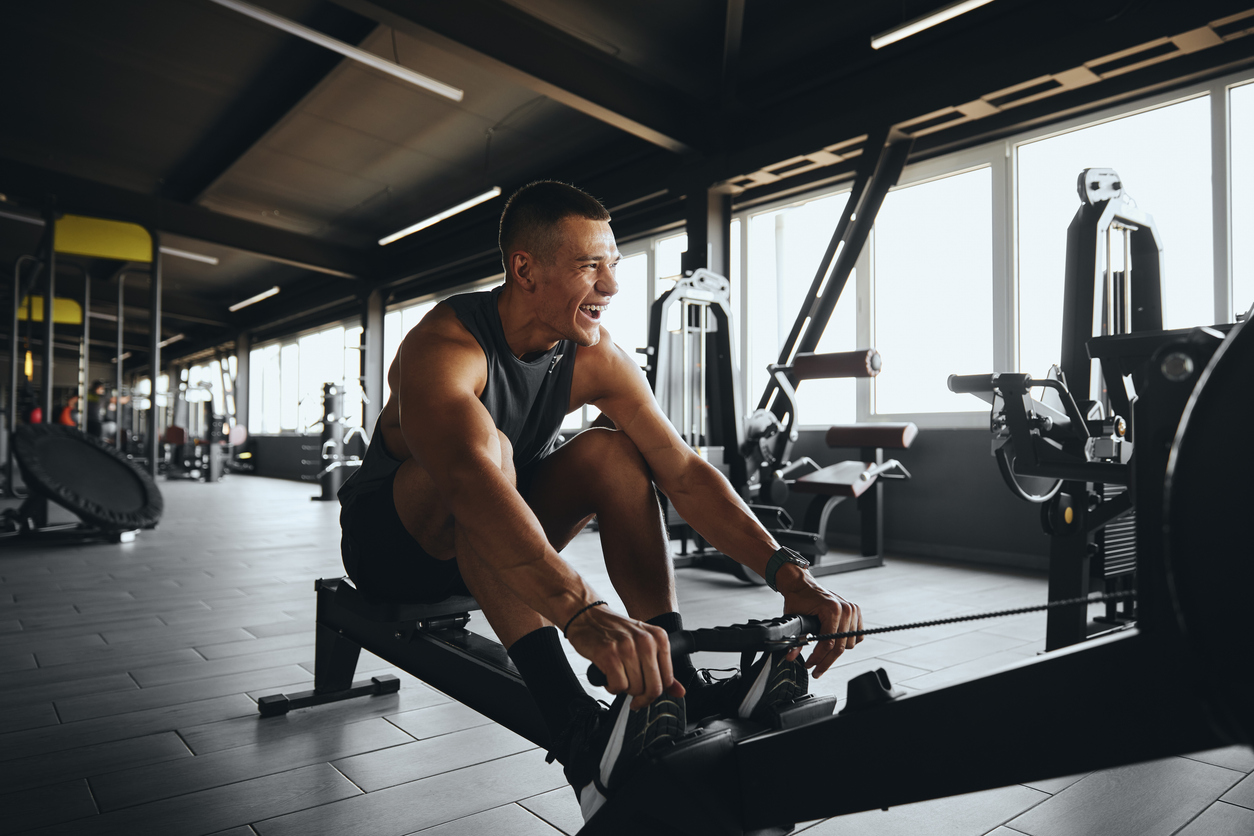 cheerful attractive man smiling while using rowing machine in contemporary gym