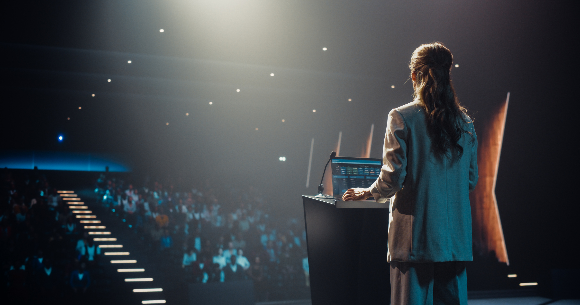 anonymous female speaker talking to the audience as she goes on conference stage. businesswoman standing with her back to camera, wearing a suit, speaking at an international business meeting