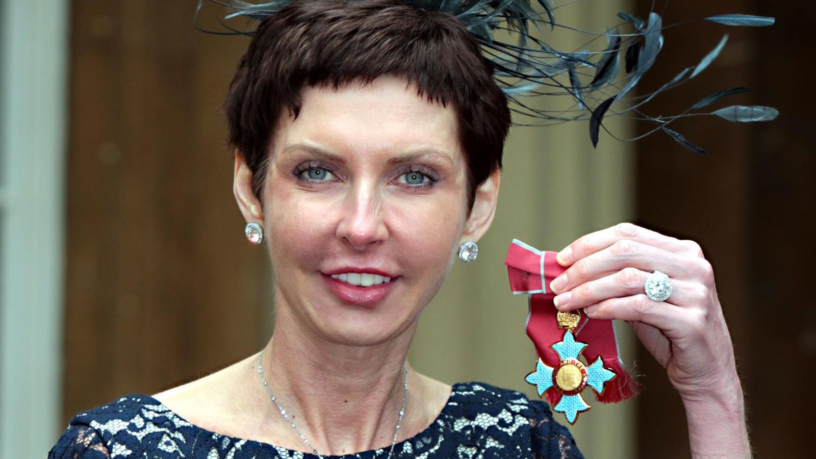 Denise Coates holding her Commander of the Order of the British Empire (CBE) medal, wearing a dark lace dress and matching earrings