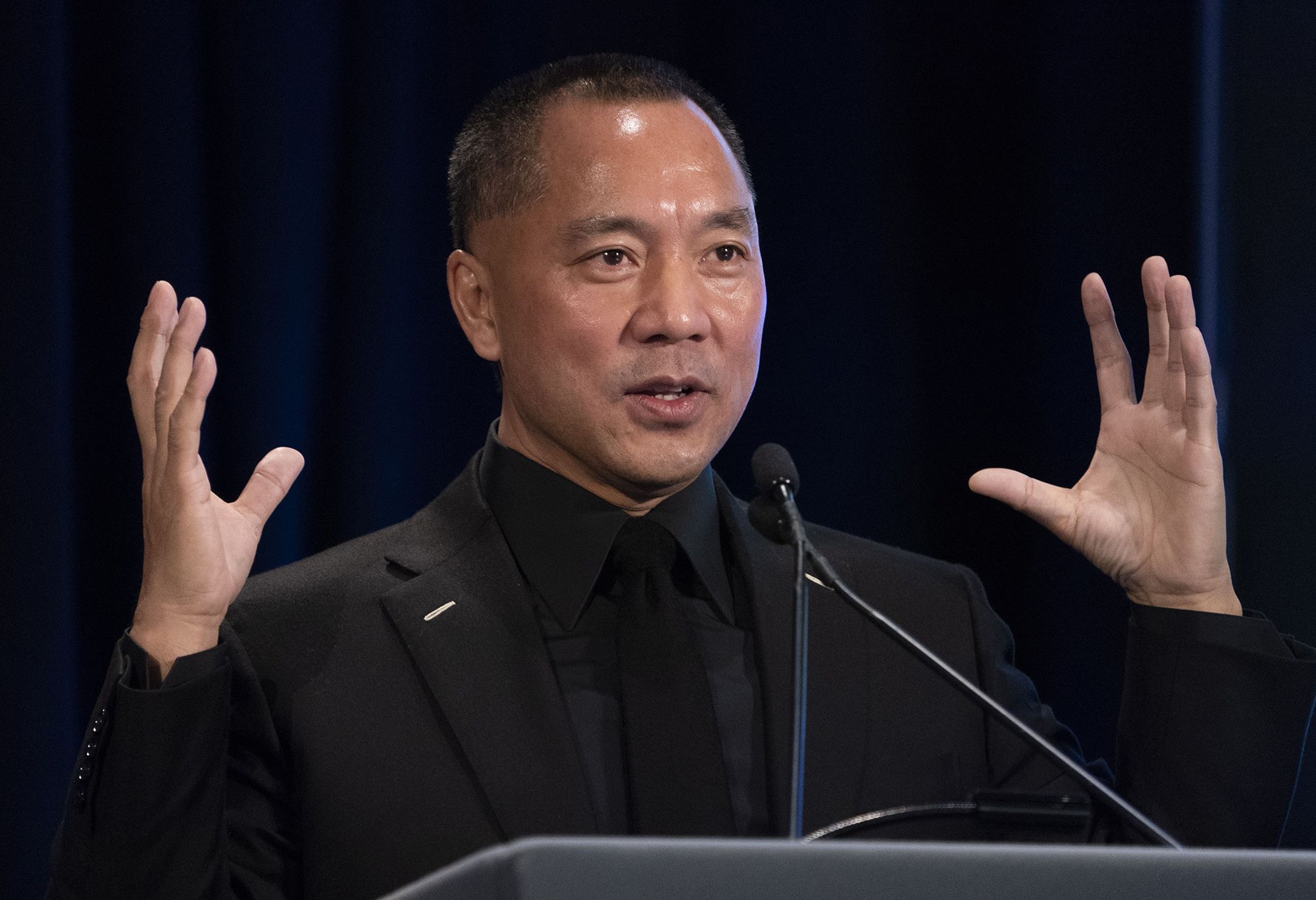 Miles Guo speaking at a public event, gesturing with both hands while standing at a podium.