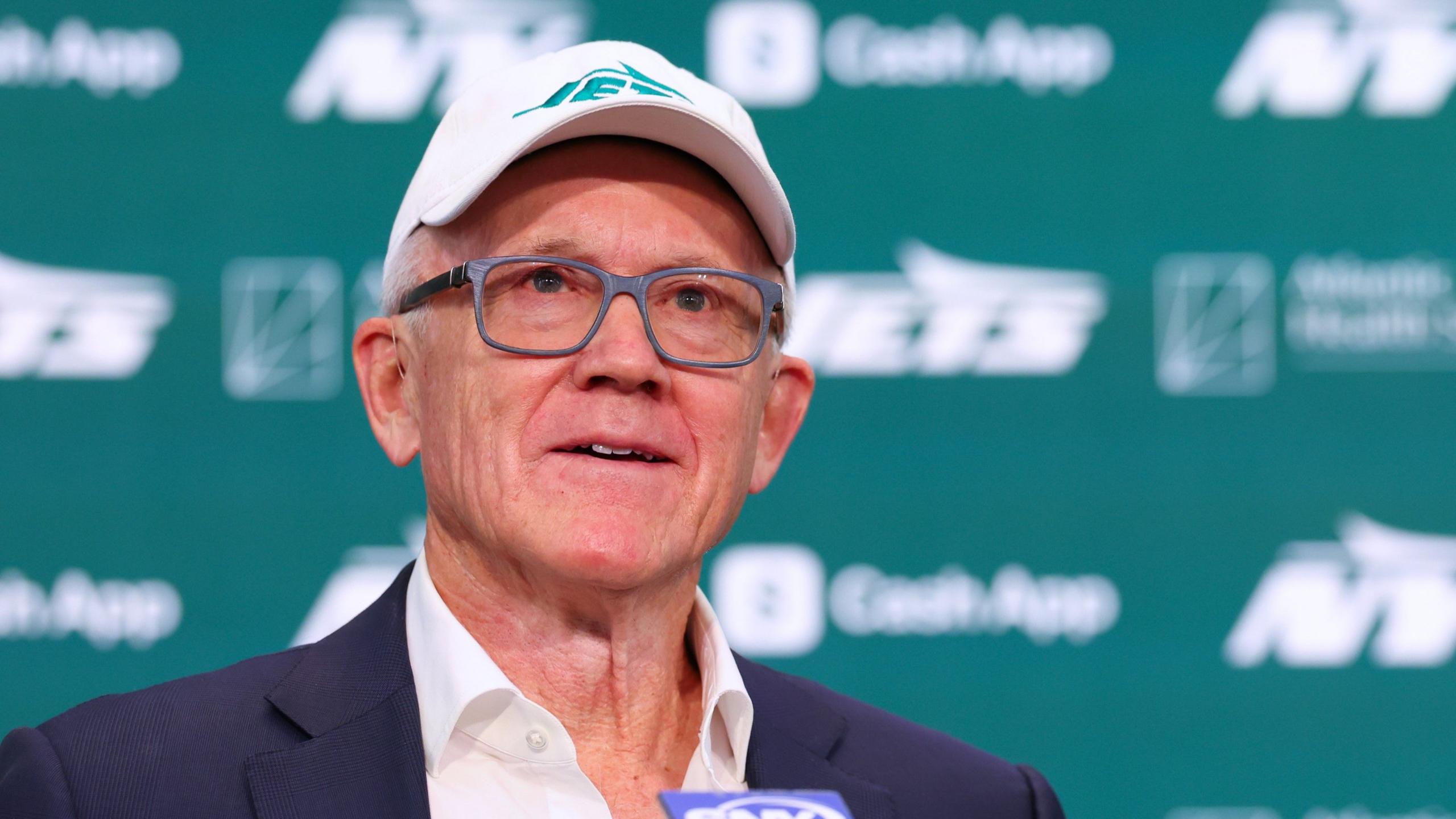 Woody Johnson wearing a New York Jets cap and glasses, speaking at a press event with a Jets-branded backdrop.