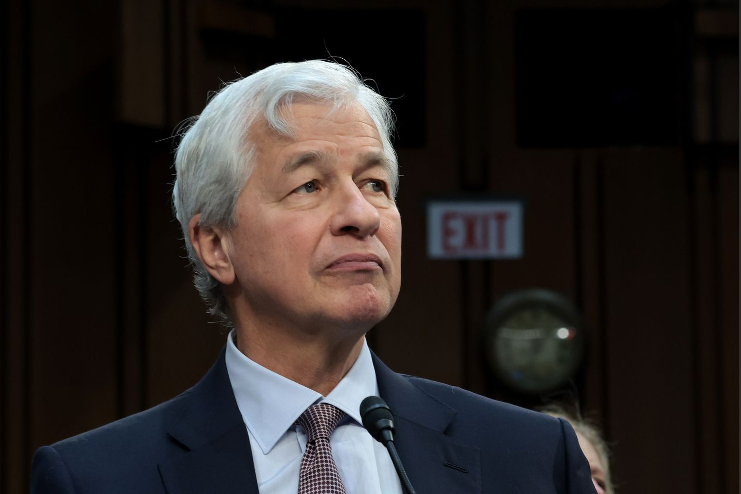 A professional portrait of Jamie Dimon, CEO of JPMorgan Chase, looking directly at the camera with a focused and serious expression. He is wearing a formal dark business suit and a blue tie, set against a neutral, minimalist office background that emphasizes executive authority.