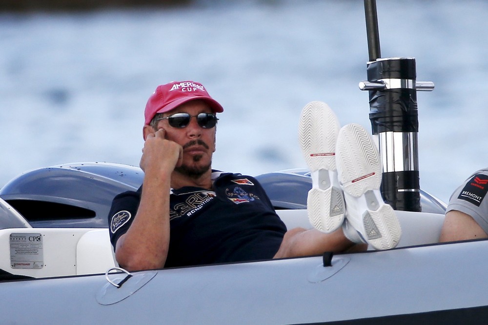 Larry Ellison relaxing aboard a luxury superyacht, wearing sunglasses and a red cap while seated on deck