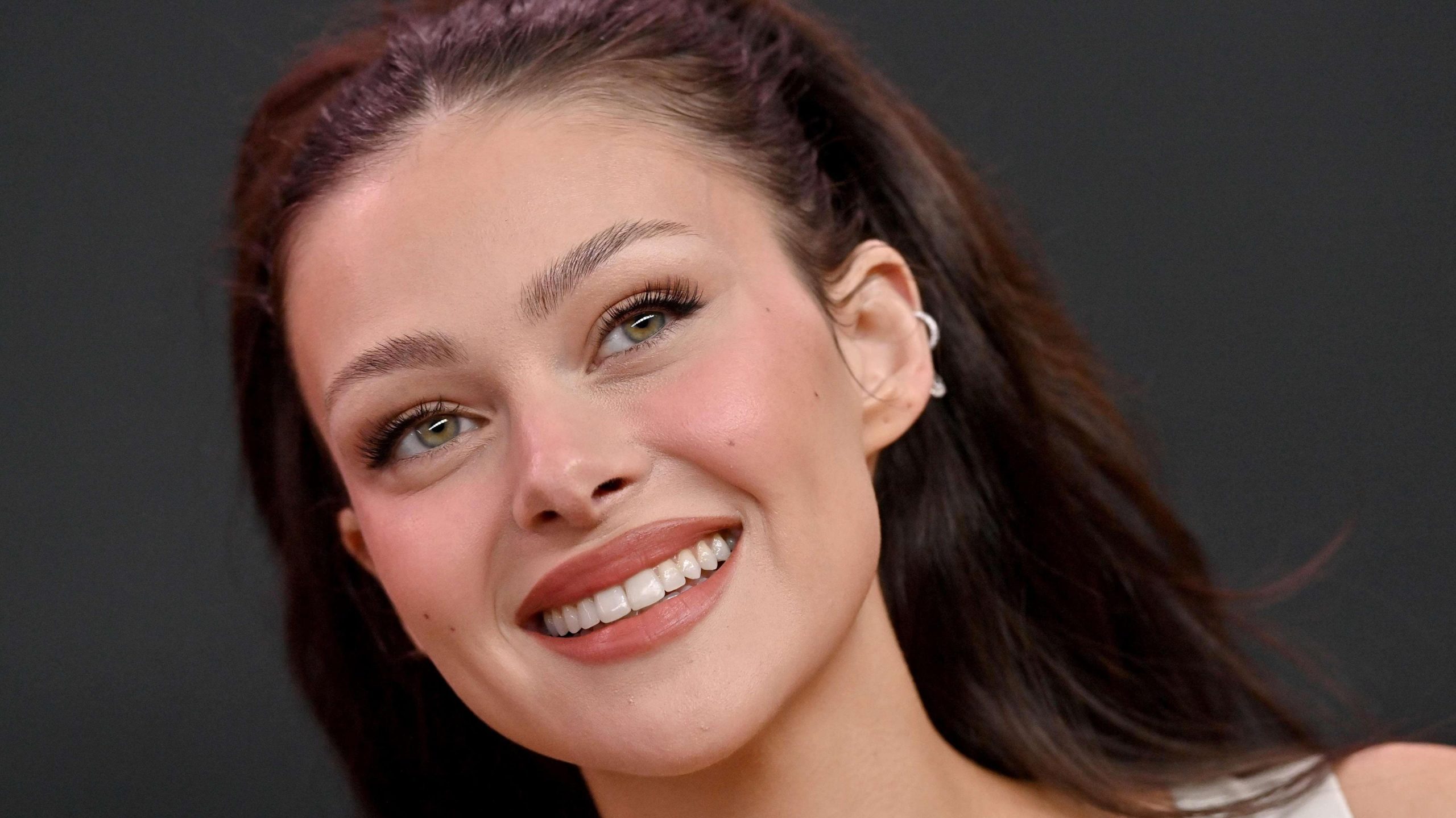 Nicola Peltz smiling at a public appearance, wearing natural makeup and a white outfit