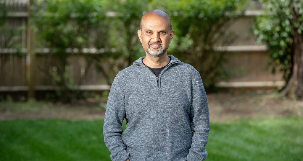 Senior business executive standing outdoors in a garden setting, wearing a grey quarter-zip sweater.
